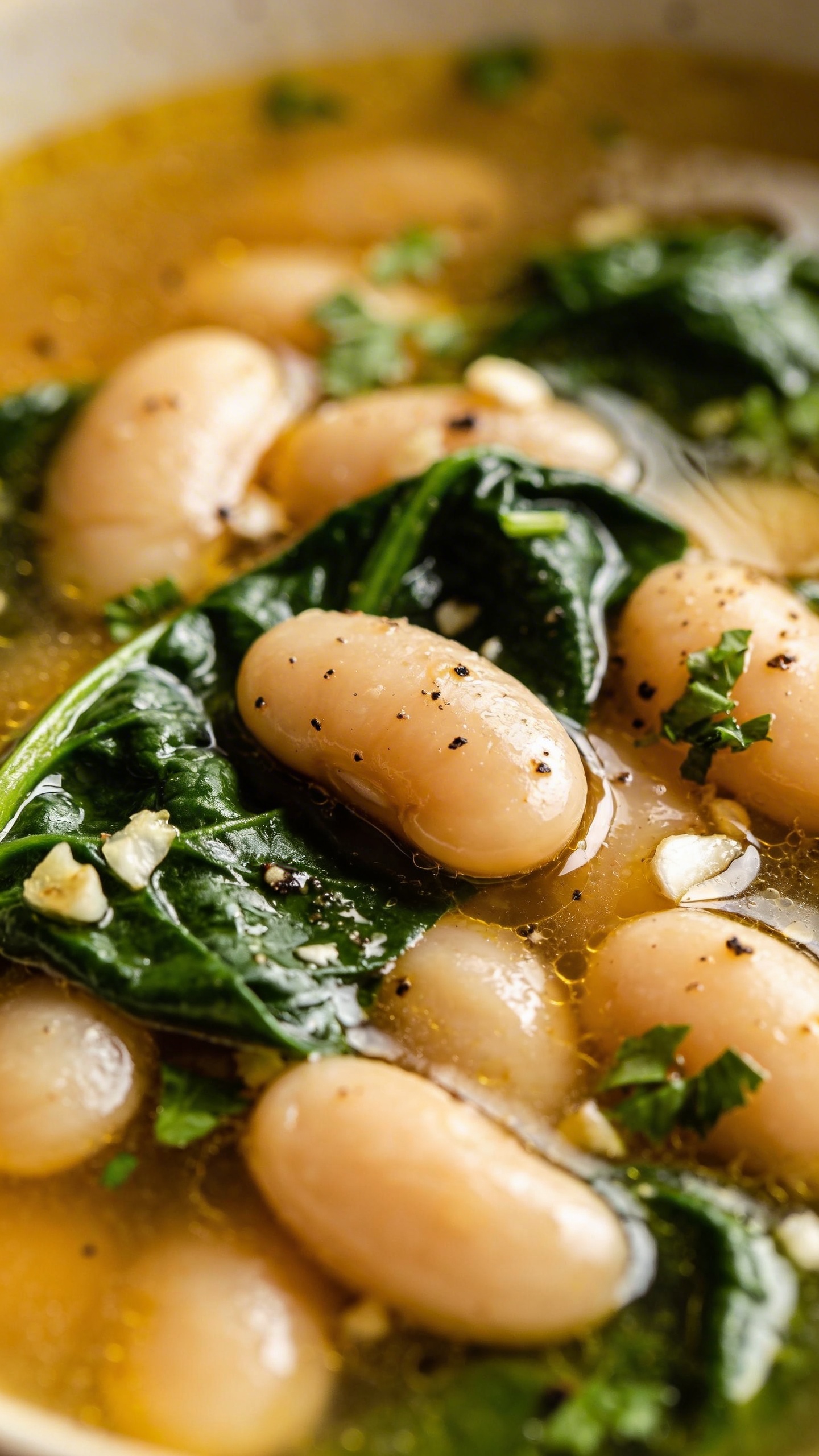 Artistic close-up of cannellini beans and fresh spinach leaves floating in golden-hued broth, with visible specs of black pepper, minced garlic, and chopped fresh parsley creating texture and depth in the composition
