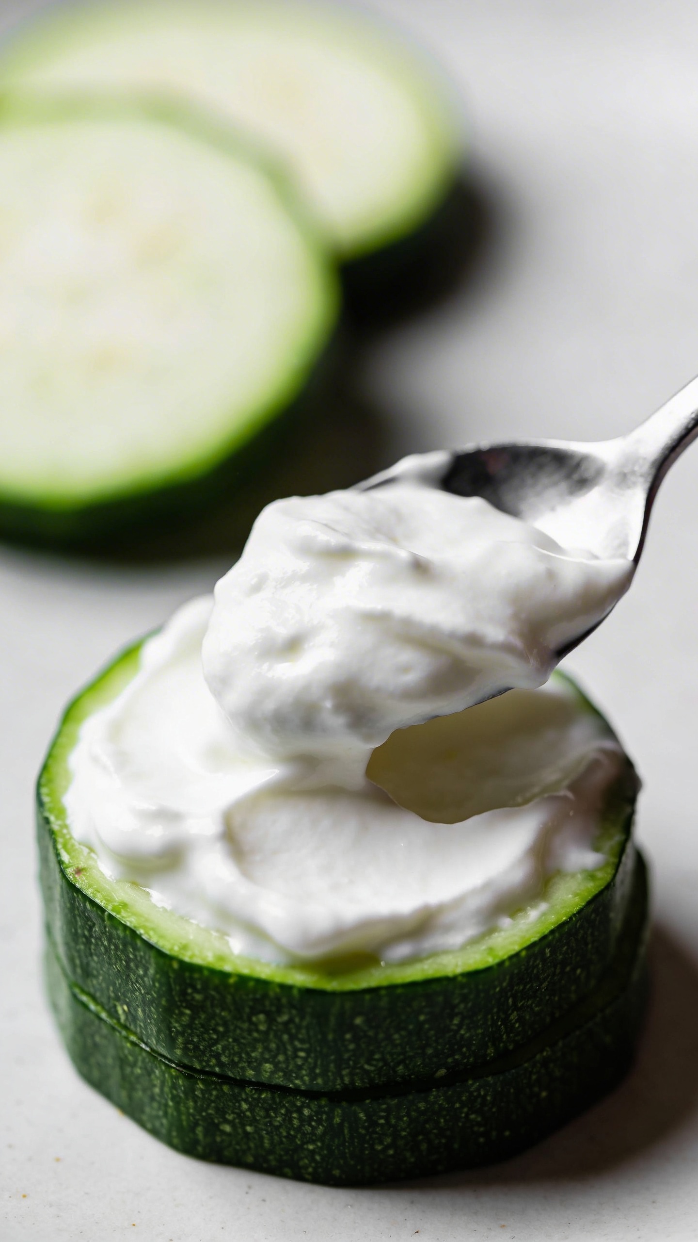 Close-up shot of thick, creamy Greek yogurt being spread onto a zucchini round with a spoon, showing the white yogurt contrasting against the green zucchini